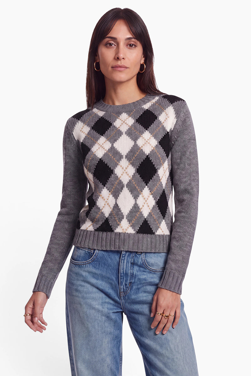 Eine Frau mit langen braunen Haaren trägt einen grauen Streick-Pullover aus einem Woll-Kaschmir-Mix mit einem Argyle-Muster aus schwarzen, weißen und beigen Rauten, dazu eine hellgewaschene Jeans. Sie steht vor einem schlichten weißen Hintergrund, blickt mit ausdruckslosem Gesicht in die Kamera und hält die Arme entspannt an den Seiten.