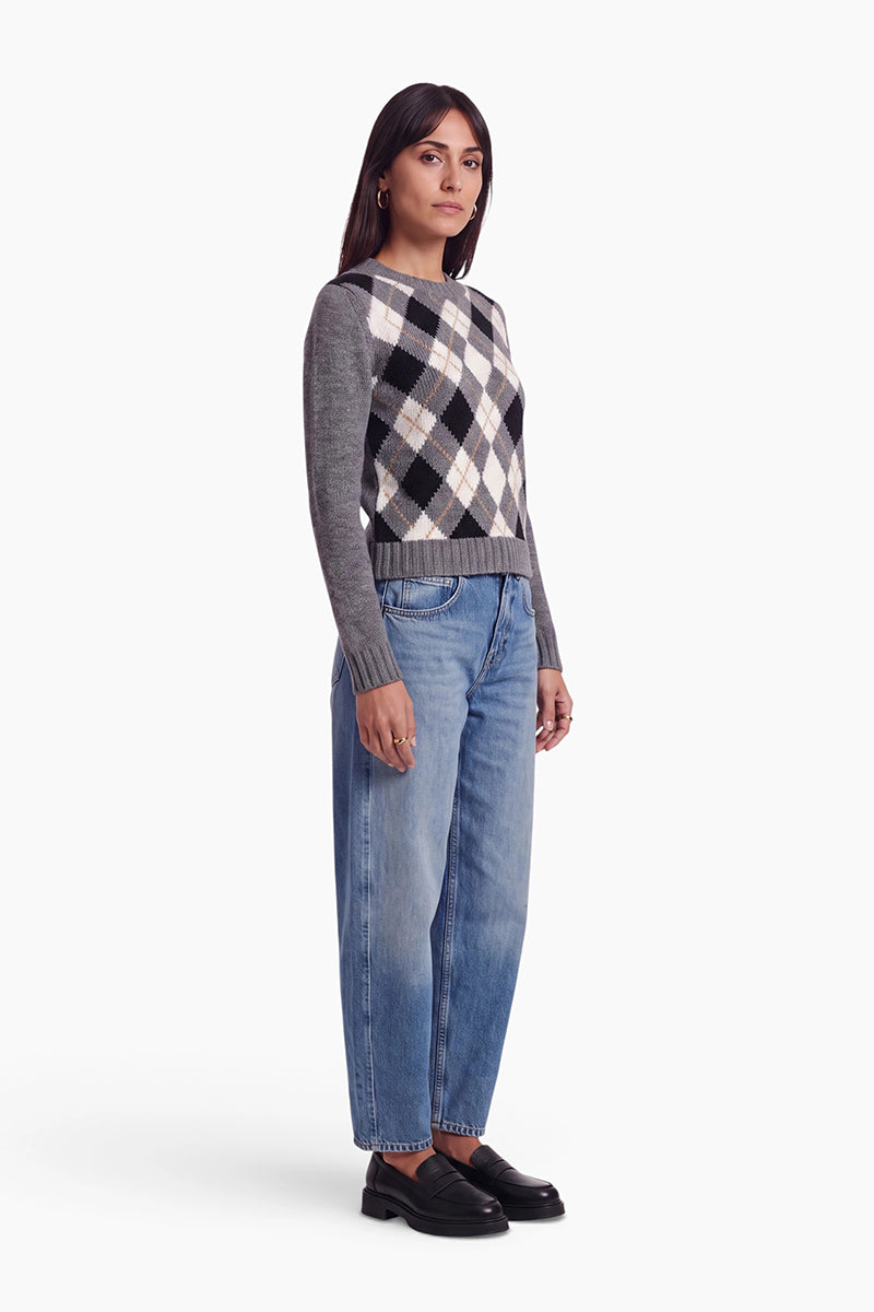 Eine Person mit langen, dunklen Haaren steht mit dem Blick nach vorne gerichtet da. Sie trägt einen grauen Pullover mit Argyle-Muster aus einer weichen Wolle-Kaschmir-Mischung, locker sitzende blaue Jeans und schwarze Slipper. Die Arme liegen locker an den Seiten, während sie vor einem schlichten weißen Hintergrund steht.