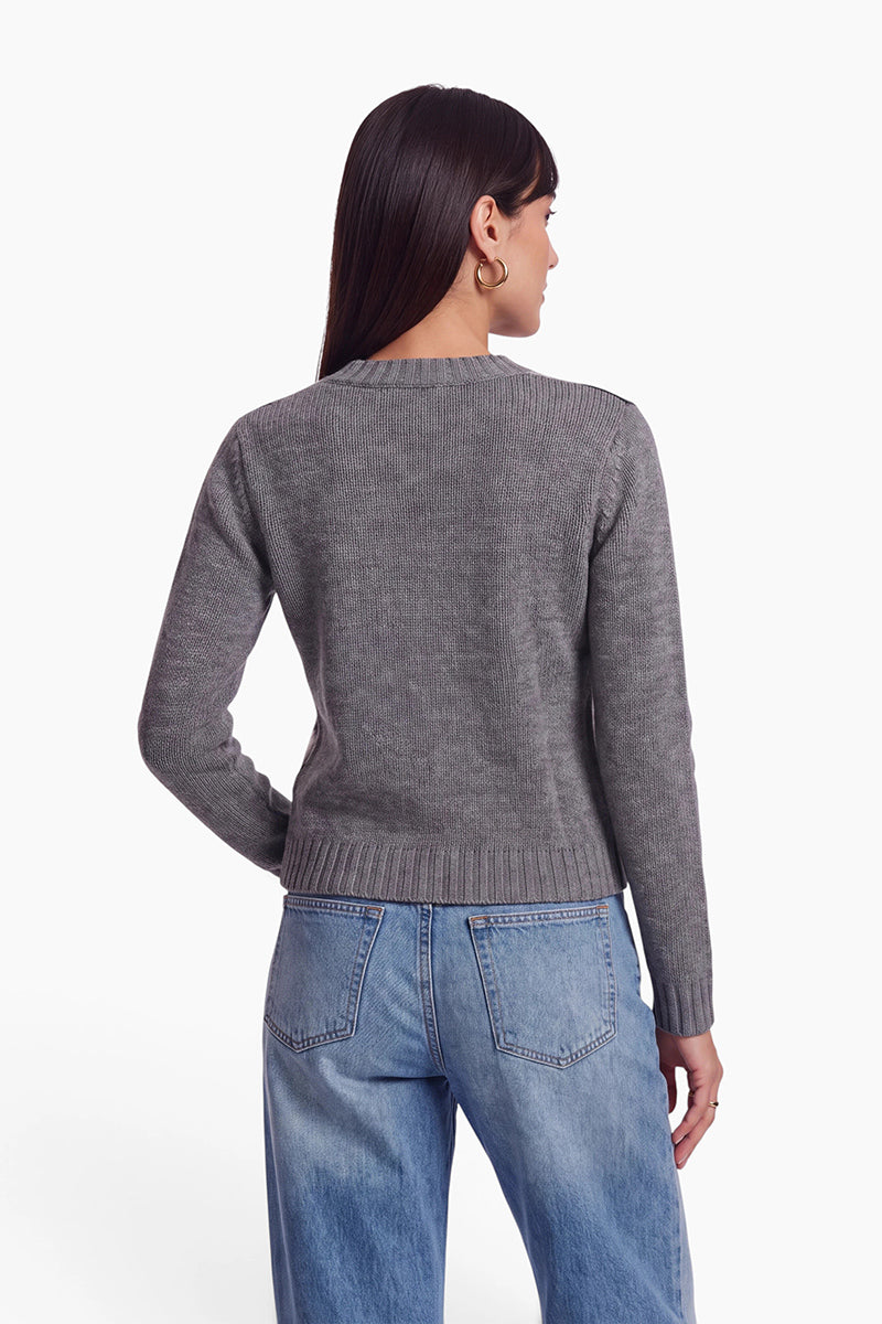 Eine Frau steht mit dem Rücken zur Kamera und trägt einen grauen Strickpullover aus einer Wolle-Kaschmir-Mischung sowie hellblaue Jeans. Ihr glattes, dunkles Haar ist seitlich gescheitelt, und sie trägt goldene Creolen. Der Hintergrund des Bildes ist schlicht weiß, wodurch die Textur und die Passform des Outfits besonders gut zur Geltung kommen.