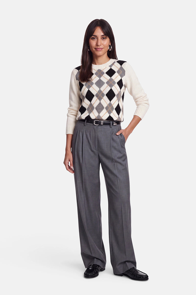 Eine Person steht vor einem weißen Hintergrund und trägt einen weißen Strickpullover aus einer Woll-Kaschmir-Mischung mit einem schwarzen, beigen und grauen Argyle-Muster, dazu eine lockere graue Bundfaltenhose, einen schwarzen Gürtel und schwarze Schuhe. Eine Hand ruht in ihrer Tasche, und sie lächelt leicht.