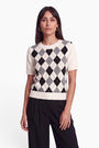 Eine Frau mit glattem braunem Haar trägt einen cremefarbenen Strickpullover mit kurzen Ärmeln, der auf der Vorderseite ein schwarz-weiß-graues Argyle-Muster aufweist und aus einer weichen Wolle-Kaschmir-Mischung gefertigt ist. Dazu trägt sie eine schwarze Hose mit hoher Taille und steht mit dem Gesicht nach vorne vor einem schlichten weißen Hintergrund.