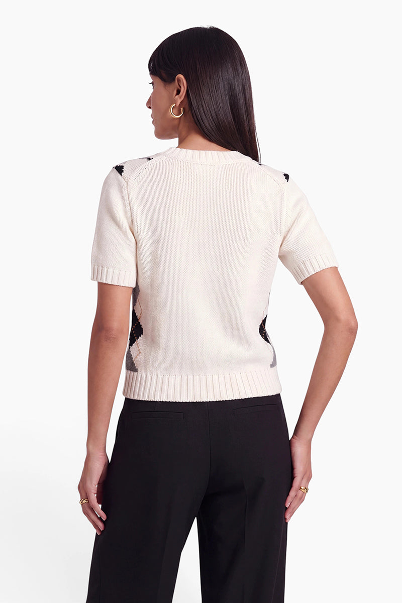 Eine Frau mit glattem, dunklem Haar trägt einen cremefarbenen Strickpullover mit kurzen Ärmeln, gerippten Bündchen und Saum sowie eine schwarze Hose. Sie ist von hinten zu sehen, wobei ein Arm angewinkelt ist und eine Hand auf ihrer Hüfte ruht. Als Accessoires trägt sie Creolen und Ringe.