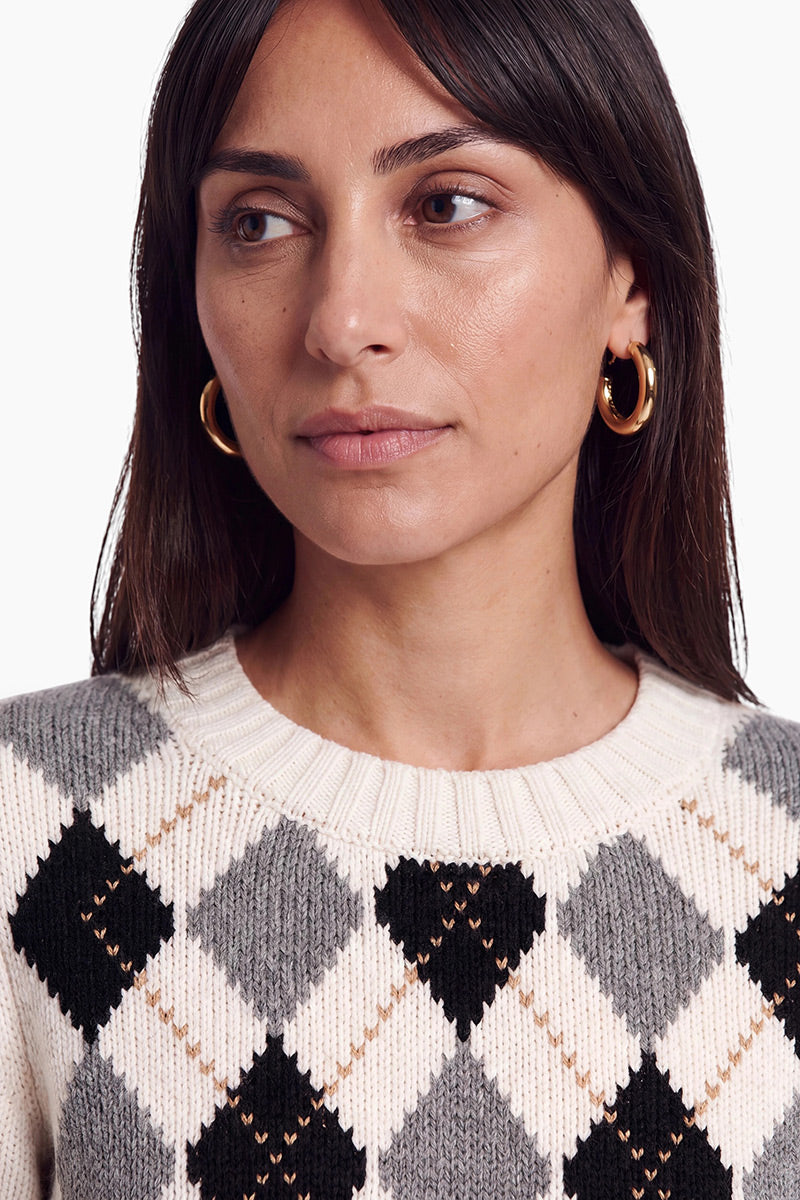 Eine Frau mit glattem braunem Haar, die goldene Creolen und einen cremefarbenen Strickpullover mit einem schwarzen, grauen und beigen Argyle-Muster trägt, blickt vor einem schlichten weißen Hintergrund leicht nach links. Der Pullover ist aus einer luxuriösen Wolle-Kaschmir-Mischung gefertigt, was ihm ein elegantes und gemütliches Aussehen verleiht.