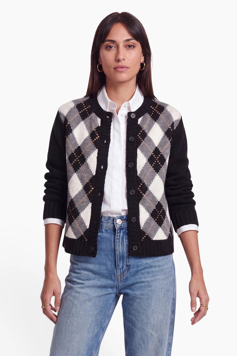 Eine Frau in hellblauen Jeans, einem weißen Hemd mit Kragen und einer schwarzen Strickjacke mit einem grau-weiß-beigen Argyle-Muster steht mit dem Blick nach vorne vor einem weißen Hintergrund. Ihre Strickjacke besteht aus einer weichen Wolle-Kaschmir-Mischung. Sie hat glattes, schulterlanges braunes Haar und einen neutralen Gesichtsausdruck.