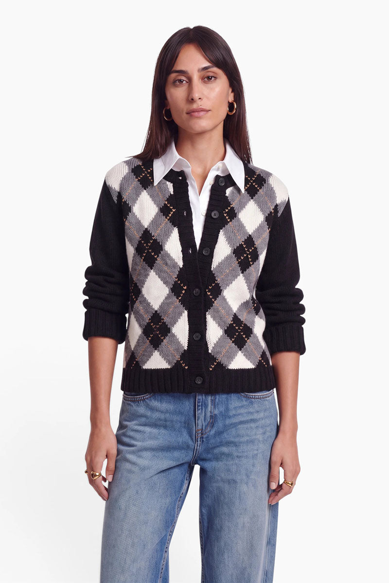 Eine Frau mit glattem braunem Haar trägt ein weißes Hemd mit Kragen unter einer schwarz-grau-weißen Strickjacke mit Argyle-Muster aus einer Woll-Kaschmir-Mischung. Außerdem trägt sie hellblaue Jeans, goldene Creolen und Ringe und steht mit neutralem Gesichtsausdruck vor einem schlichten weißen Hintergrund.