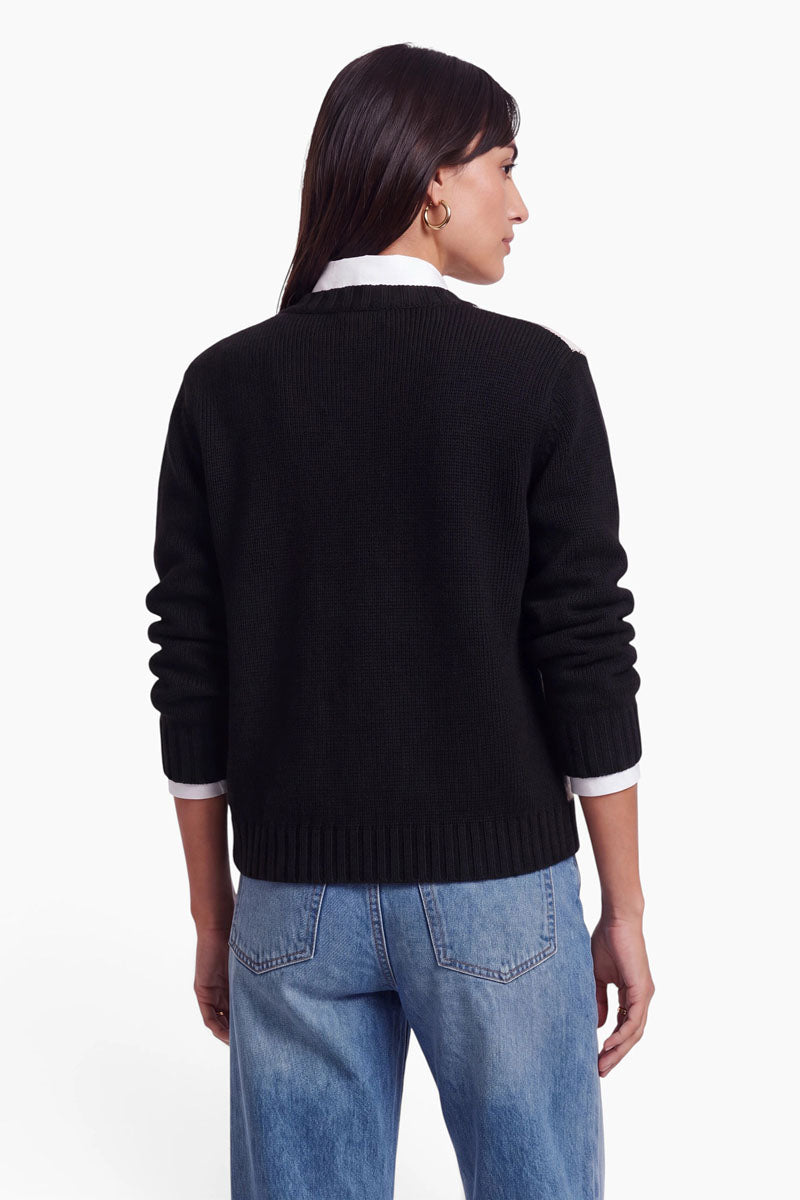 Eine Person mit glattem, dunklem Haar, die eine schwarze Strickjacke aus einer Wolle-Kaschmir-Mischung über einem weißen Hemd mit Kragen und blauen Jeans trägt, steht mit dem Rücken zur Kamera vor einem schlichten weißen Hintergrund.