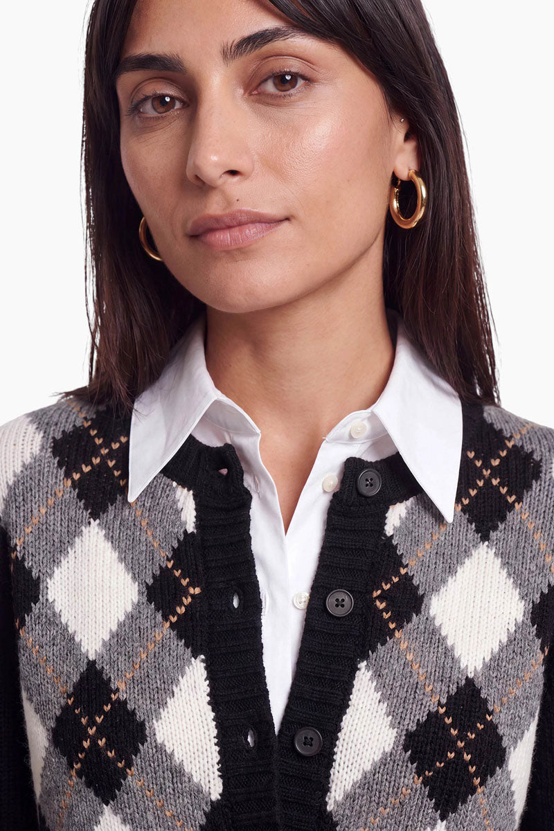 Eine Frau mit glattem, dunklem Haar trägt goldene Creolen, ein weißes Hemd mit Kragen und eine schwarz-grau-weiße Strickjacke mit Argyle-Muster aus einer Woll-Kaschmir-Mischung. Mit ausdruckslosem Gesicht blickt sie leicht an der Kamera vorbei. Der Hintergrund ist schlicht weiß.