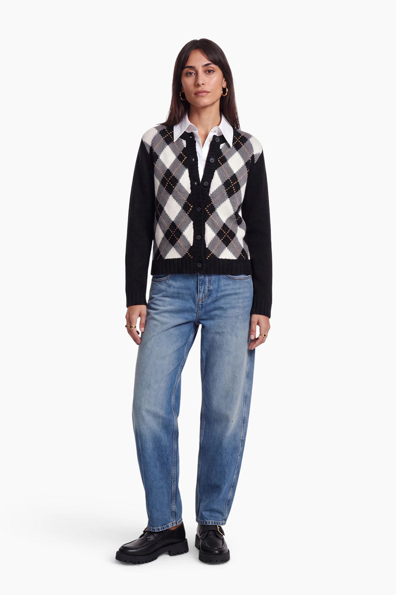 Eine Frau steht mit dem Blick nach vorne und trägt eine Strickjacke aus einem Woll-Kaschmir-Mix mit einem klassischen Argyle-Muster in Schwarz, Weiß und Grau über einem weißen Hemd mit Kragen, hellblauen Jeans und schwarzen, klobigen Schuhen. Sie hat glattes, dunkles Haar, eine entspannte Körperhaltung und einen neutralen Gesichtsausdruck. Der Hintergrund ist schlicht weiß.