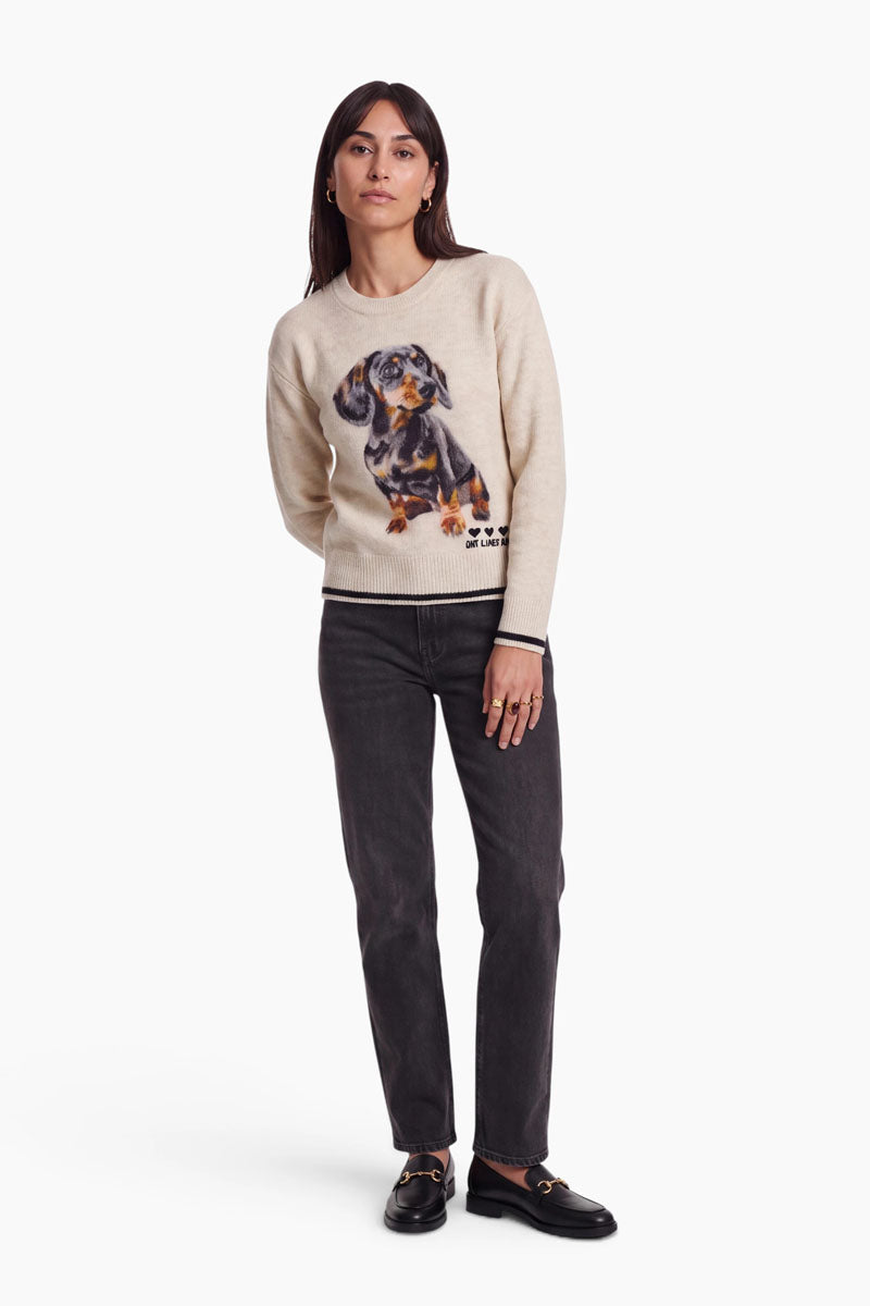 Eine Frau mit langen dunklen Haaren steht mit dem Blick nach vorne. Sie trägt einen beigen Damen-Strickpullover mit einem Dackelmotiv, schwarze Jeans und schwarze Slipper. Der kuschelige Pullover besticht durch einen klassischen Rundhalsausschnitt, der ihm einen zeitlosen Charme verleiht. Sie trägt Creolen und Ringe und hat eine Hand in der Tasche. Der Hintergrund ist komplett weiß.
