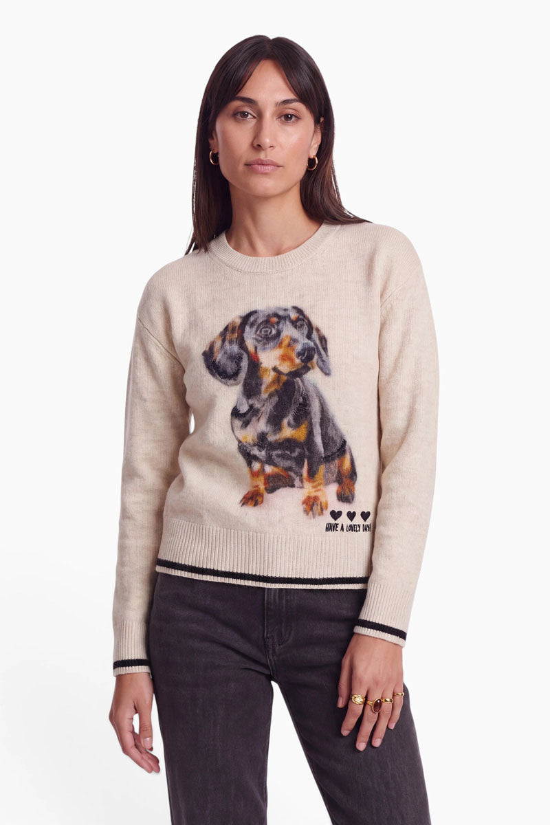 Eine Frau mit glattem braunem Haar trägt einen cremefarbenen Strickpullover für Damen mit einem realistischen Dackel-Motiv und kleinen schwarzen Herzen, dazu eine dunkle Jeans. Der gemütliche Pullover mit klassischem Rundhalsausschnitt hebt sich deutlich von ihrem neutralen Gesichtsausdruck ab, während sie vor einem weißen Hintergrund in die Kamera blickt.