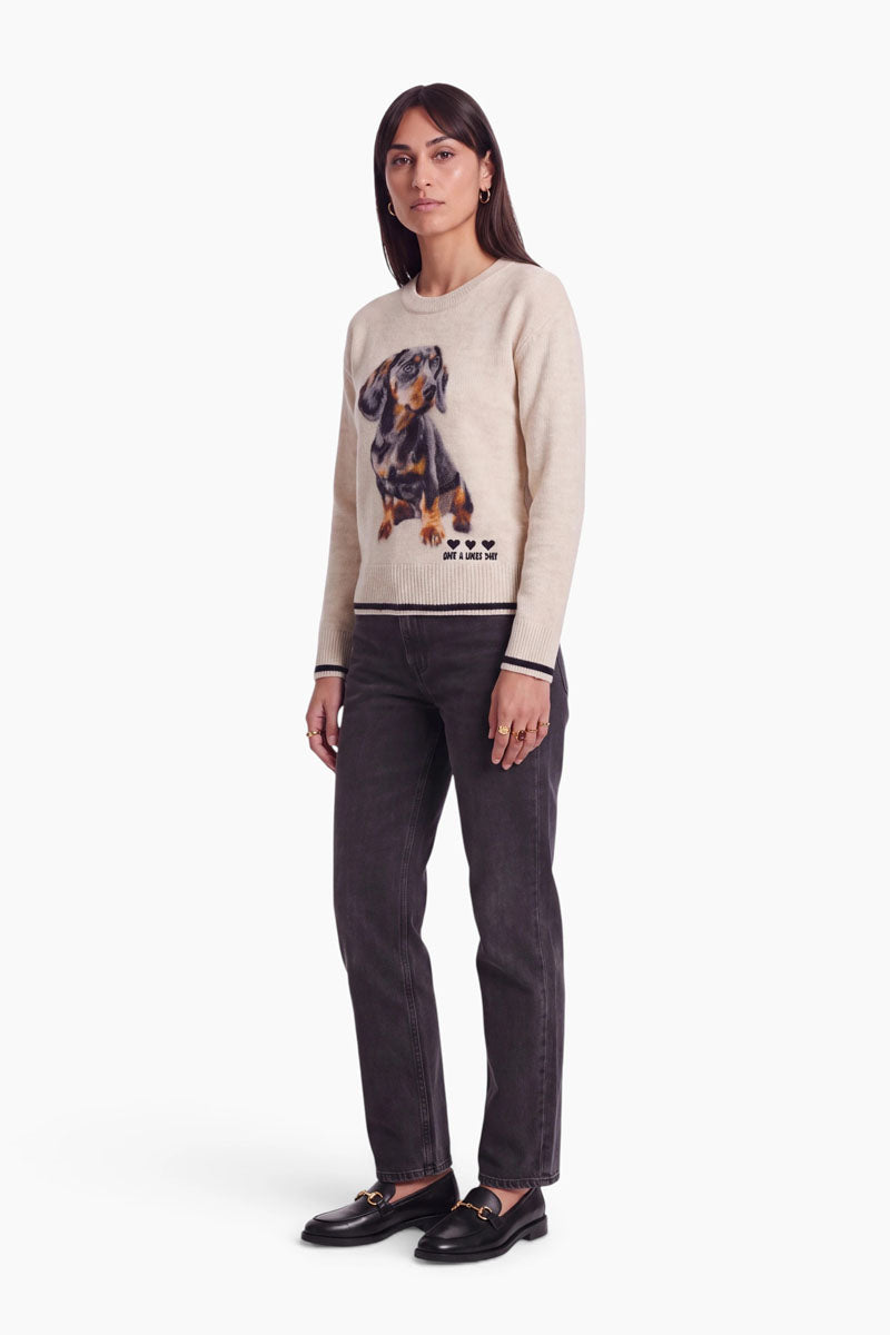 Eine Frau mit glattem braunem Haar steht vor einem schlichten weißen Hintergrund und trägt einen beigen Damen-Strickpullover mit Rundhalsausschnitt und einem Dackel-Motiv, dunkelgraue Jeans und schwarze Loafer. Sie blickt mit neutralem Gesichtsausdruck geradeaus.