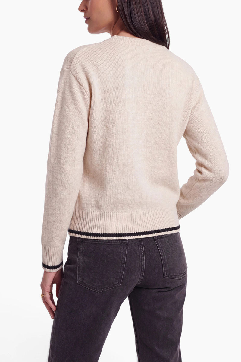Eine Person mit langen braunen Haaren trägt einen cremefarbenen Pullover aus Alpaka-Wolle mit schwarz-weißen Bordüren an den Ärmelbündchen und am Saum, dazu eine dunkelgraue Jeans. Der Damen-Strickpullover verfügt über einen klassischen Rundhalsausschnitt. Die Person steht mit dem Rücken zur Kamera vor einem weißen Hintergrund.