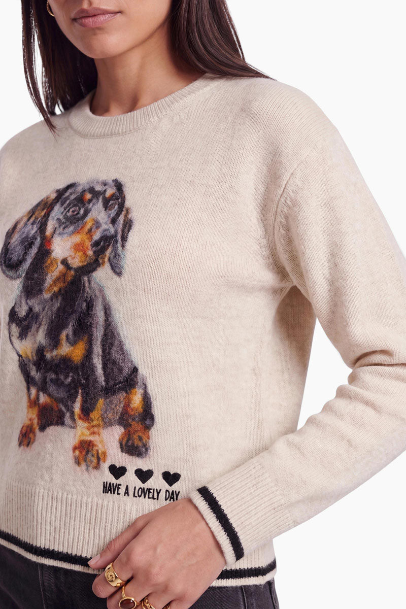 Eine Frau trägt einen cremefarbenen Strickpullover für Damen mit einer großen, realistischen Dackel-Illustration und dem Aufdruck „HAVE A LOVELY DAY“ sowie drei schwarzen Herzen nahe dem Saum. Der kuschelige Pullover aus Alpaka-Wolle mit klassischem Rundhalsausschnitt wird zu einer dunklen Hose und goldenen Ringen kombiniert. Nur ihr unterer Gesichtsbereich und ihr Oberkörper sind zu sehen.