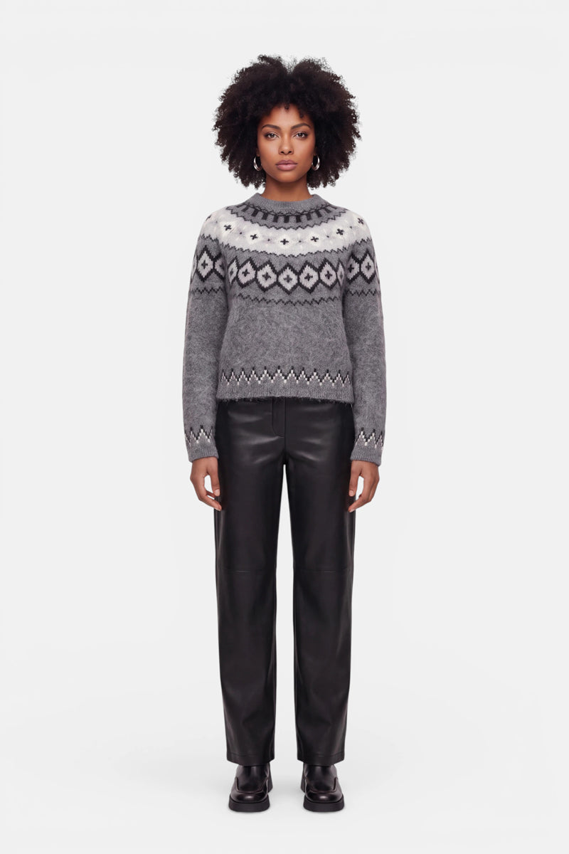 Eine Frau steht mit dem Blick nach vorne und trägt einen grauen Strickpullover aus einer Woll-Kaschmir-Mischung mit einem auffälligen Jacquard-Muster in Schwarz und Weiß, dazu eine schwarze Lederhose und schwarze Stiefel. Sie hat lockiges Haar und einen neutralen Gesichtsausdruck und posiert vor einem schlichten weißen Hintergrund.