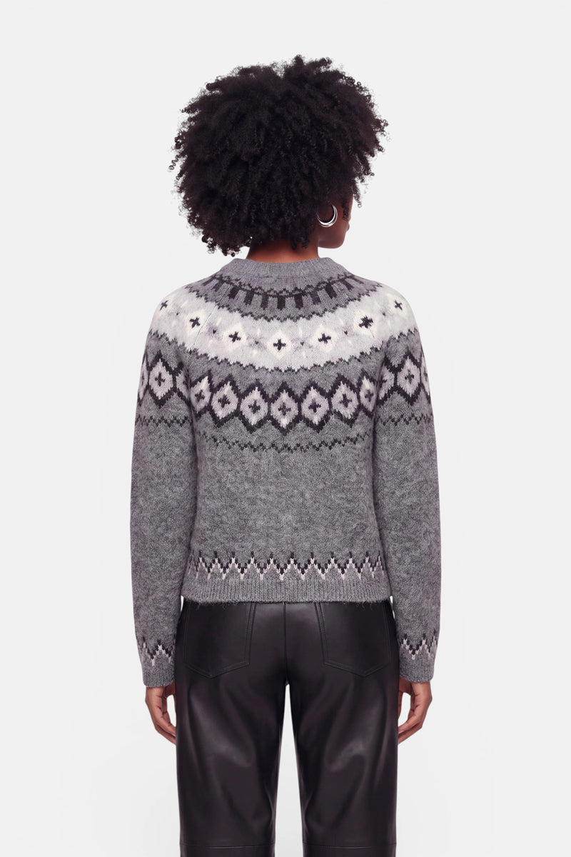 Eine Person mit kurzen, lockigen Haaren trägt einen grauen Strickpullover mit einem geometrischen, schwarz-weißen Jacquard-Muster im Schulterbereich sowie eine schwarze Lederhose. Die Person steht mit dem Rücken zur Kamera vor einem schlichten weißen Hintergrund.