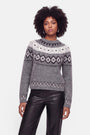 Eine Frau mit lockigem Haar trägt Creolen, einen grauen Strickpullover mit weiß-schwarzem Jacquard-Muster und eine schwarze Lederhose. Der Strickpullover besteht aus einer edlen Wolle-Kaschmir-Mischung. Sie steht vor einem schlichten weißen Hintergrund, blickt nach vorne und hat einen neutralen Gesichtsausdruck.
