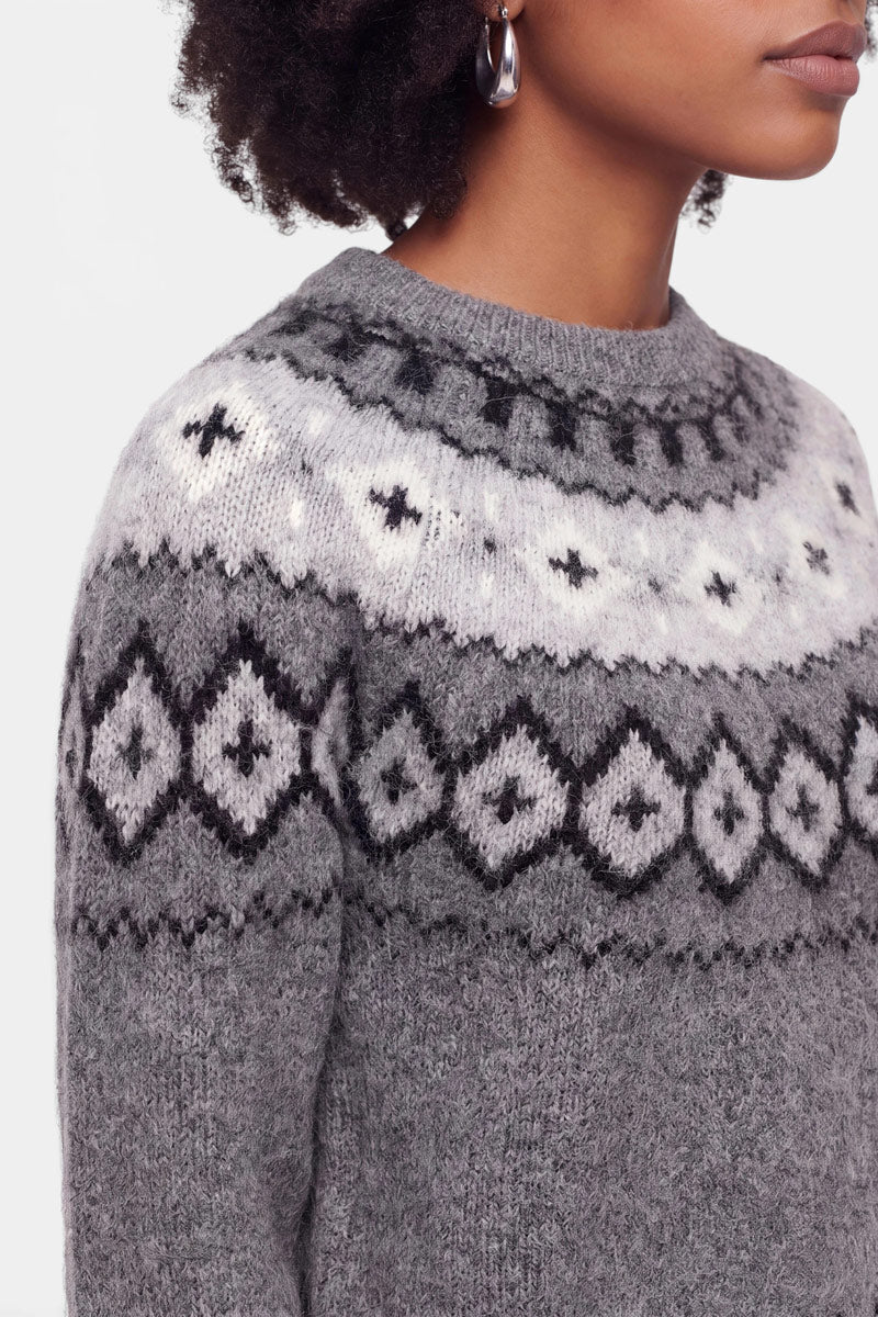Eine Person, die einen grauen Strickpullover mit einem weiß-schwarzen Jacquard-Muster über Brust und Schultern trägt. Zu sehen sind nur der untere Teil des Gesichts, der Hals und der obere Oberkörper. Die Person hat lockiges Haar und trägt silberne Creolen. Der Pullover ist aus einer weichen Wolle-Kaschmir-Mischung gefertigt und bietet somit zusätzlichen Tragekomfort.