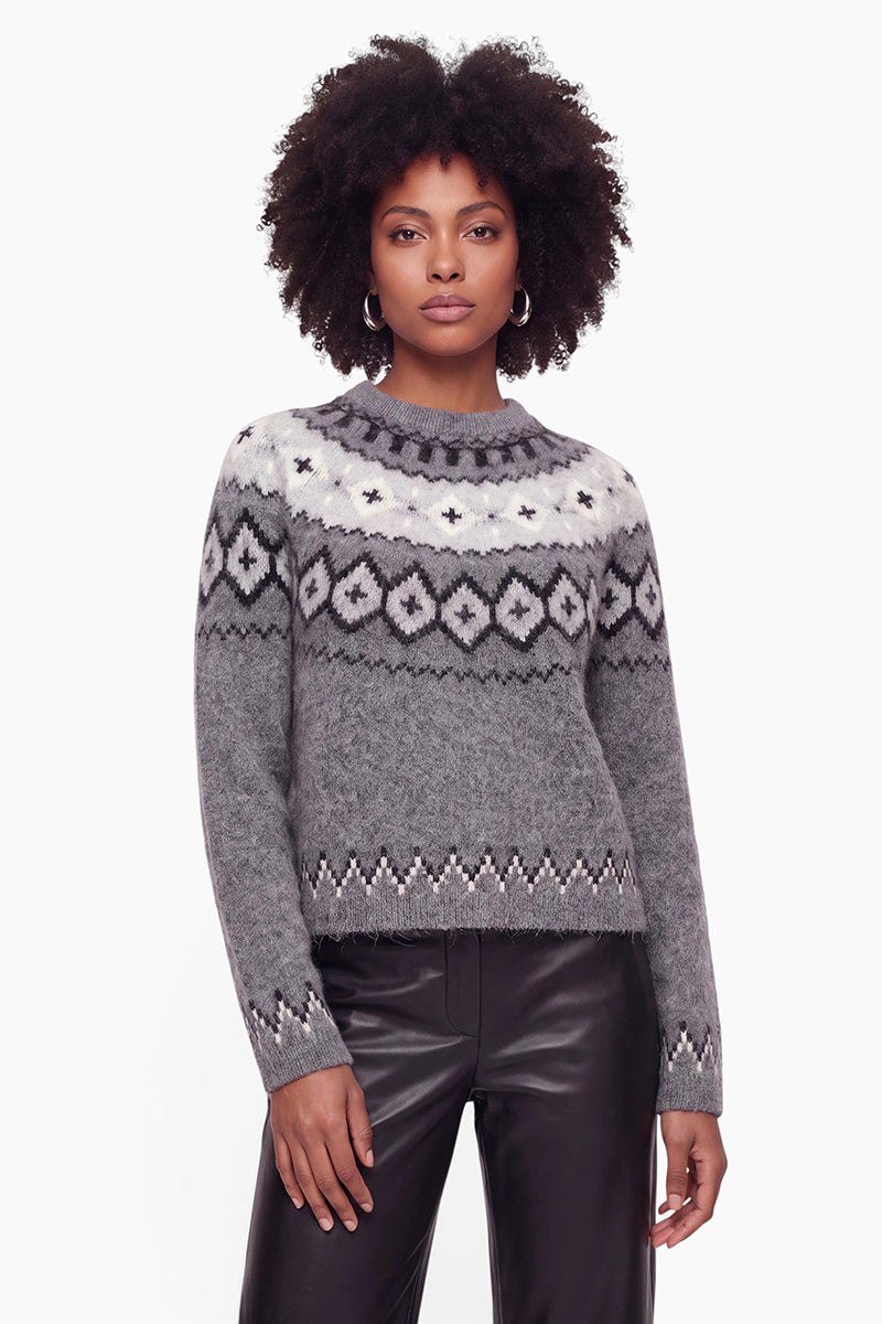 Eine Frau mit lockigem Haar trägt einen grauen, gemusterten Strickpullover aus gebürsteter Wolle mit weißen und schwarzen Jacquard-Mustern an Passe und Ärmelbündchen, dazu eine schwarze Lederhose. Sie steht vor einem schlichten weißen Hintergrund und blickt selbstbewusst in die Kamera.