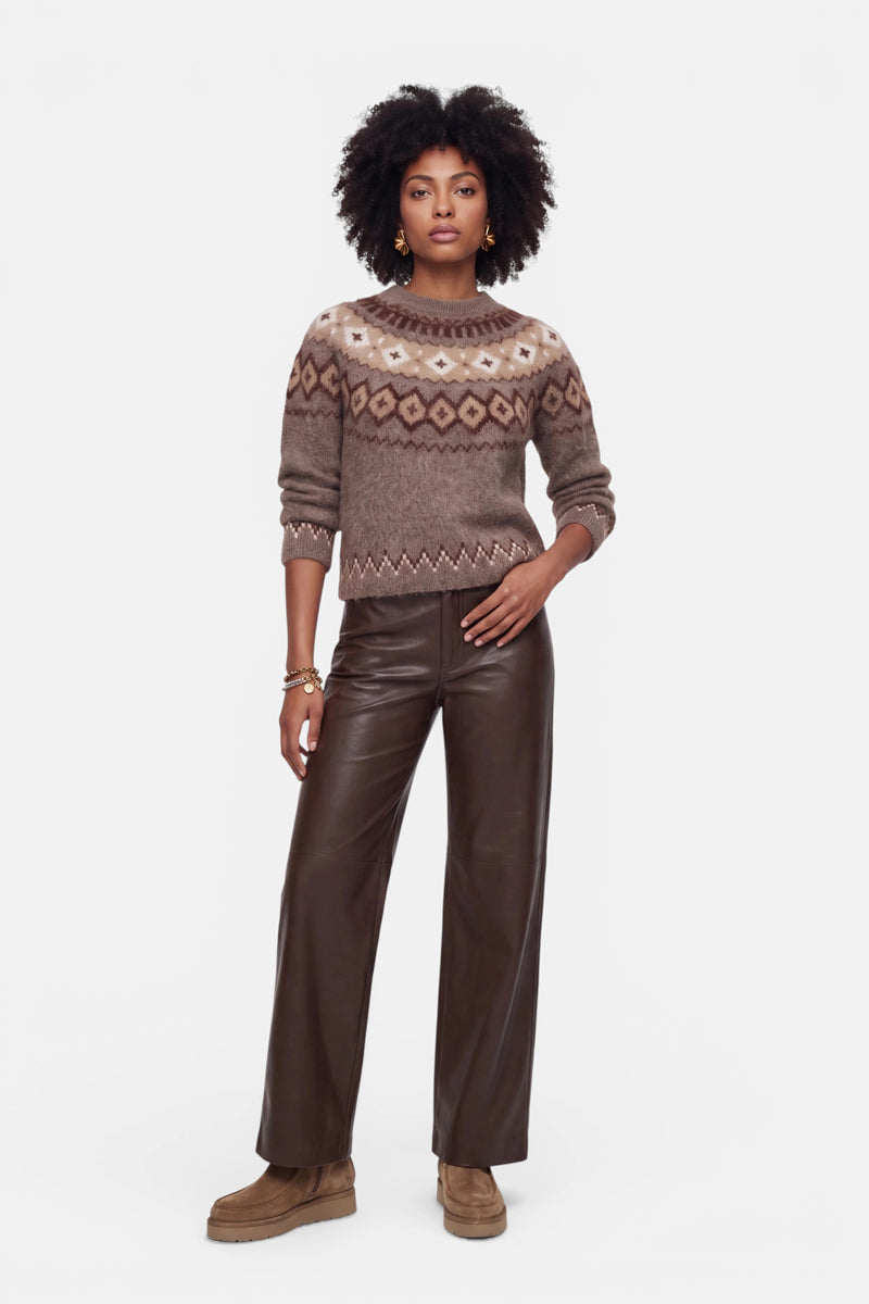 Eine Frau mit natürlich lockigem Haar trägt einen braunen Strickpullover mit Jacquard-Muster, eine dunkelbraune Lederhose mit weitem Bein und braune Schuhe. Sie steht mit einer Hand in der Tasche vor einem schlichten weißen Hintergrund und blickt geradeaus.
