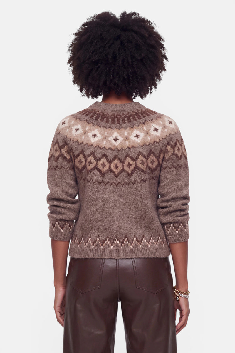 Eine Person mit lockigem, dunklem Haar ist von hinten zu sehen. Sie trägt einen braun gemusterten Strickpullover aus einem Woll-Kaschmir-Mix mit geometrischen Jacquard-Mustern in Beige und Creme sowie eine dunkelbraune Lederhose. Die Arme liegen entspannt an den Seiten, und an einem Handgelenk trägt sie Armbänder. Der Hintergrund ist einfarbig weiß.