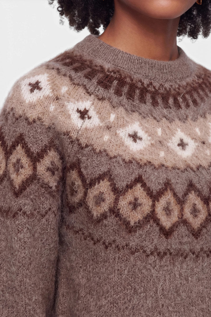 Eine Person, die einen flauschigen braunen Strickpullover aus einer Woll-Kaschmir-Mischung trägt, der auf Brust und Schultern ein Jacquard-Muster aus Rauten und Sternen in Creme und Dunkelbraun aufweist. Nur der untere Teil des Gesichts, der Hals und der obere Oberkörper sind zu sehen. Der Hintergrund ist einfarbig weiß.