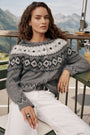 Eine Frau mit langen, welligen braunen Haaren trägt eine grau-weiße Strickjacke mit einem eleganten Jacquard-Muster, die aus einer weichen Woll-Kaschmir-Mischung gefertigt ist. Sie sitzt draußen auf einem Holzstuhl, im Hintergrund sind Berge und Grünflächen zu sehen, und blickt selbstbewusst in die Kamera.
