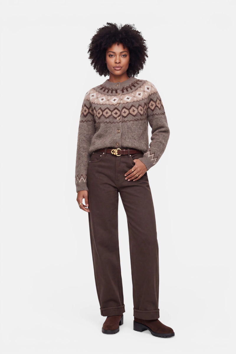 Eine Frau mit lockigem Haar trägt einen braun-beigen Pullover mit Jacquard-Muster aus einer luxuriösen Wolle-Kaschmir-Mischung, dazu eine braune Hose mit weitem Bein, einen braunen Gürtel mit goldener Schnalle und braune Schuhe. Sie steht mit dem Gesicht nach vorne vor einem schlichten weißen Hintergrund.