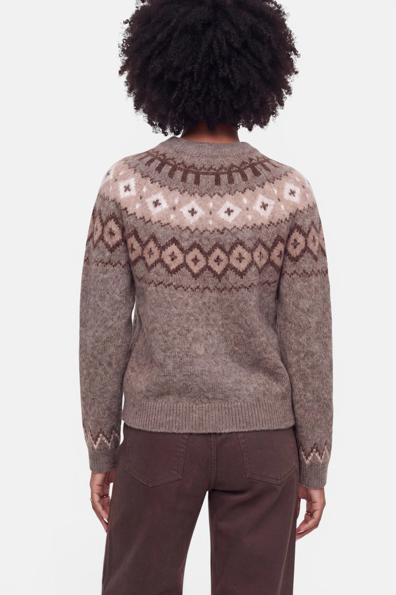 Eine Person mit lockigem Haar, von hinten gesehen, trägt eine braune Fair-Isle-Strickjacke aus einer luxuriösen Wolle-Kaschmir-Mischung mit geometrischen Jacquard-Mustern in Beige, Hellbraun und Creme auf dem Passe und den Ärmeln, dazu eine dunkelbraune Hose. Der Hintergrund ist einfarbig weiß.
