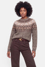 Eine Frau mit natürlich lockigem Haar trägt eine taupefarbene Strickjacke aus einer Woll-Kaschmir-Mischung mit Fair-Isle-Jacquard-Muster in Beige und Braun, die sie in eine dunkelbraune Hose mit hoher Taille gesteckt hat. Sie steht mit dem Blick nach vorne, die Hände entspannt, und einem neutralen Gesichtsausdruck vor einem schlichten weißen Hintergrund.