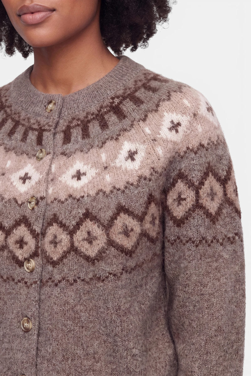 Eine Frau trägt eine braun-beige Strickjacke aus einer edlen Wolle-Kaschmir-Mischung, die über Brust und Schultern mit einem geometrischen Fair-Isle-Jacquard-Muster verziert ist. Das Kleidungsstück hat einen Rundhalsausschnitt und wird mit fünf braunen Knöpfen geschlossen. Zu sehen sind nur ihr unterer Gesichtsbereich, ihr Hals und ihr oberer Oberkörper.