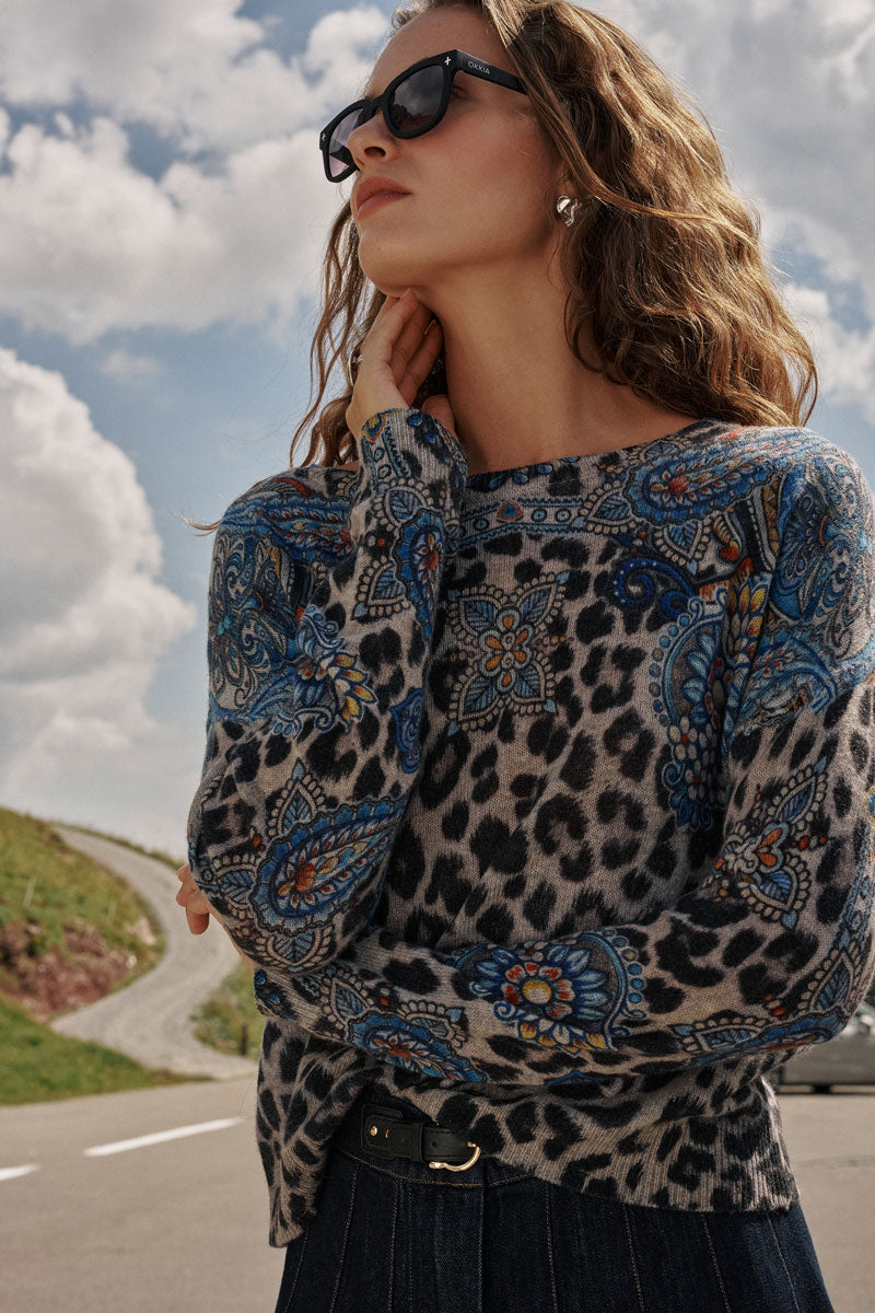 Eine Frau mit welligem braunem Haar und Sonnenbrille steht im Freien vor einem bewölkten Himmel. Sie trägt einen stilvollen Feinstrickpullover mit einem Leo-Paisley-Muster in blauen Blumenmotiven, der an der Taille mit einem schwarzen Gürtel gerafft ist und zu einem dunklen Faltenrock kombiniert wird. Im Hintergrund sind eine kurvenreiche Straße und ein grasbewachsener Hang zu sehen, die ihren schicken Look unterstreichen.