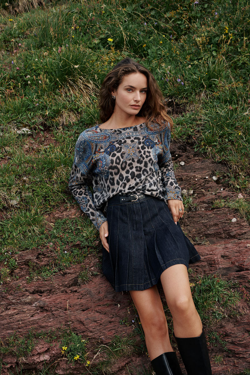 Eine Frau mit welligem braunem Haar liegt auf einem grasbewachsenen, felsigen Hang. Sie trägt einen langärmeligen, gemusterten Pullover mit Tier- und Paisley-Motiven sowie einen plissierten Mini-Jeansrock in dunkler Waschung mit einem Gürtel. Ihre schwarzen kniehohen Stiefel runden den Look ab, während sie von Wildblumen und grünem Gras umgeben ist.
