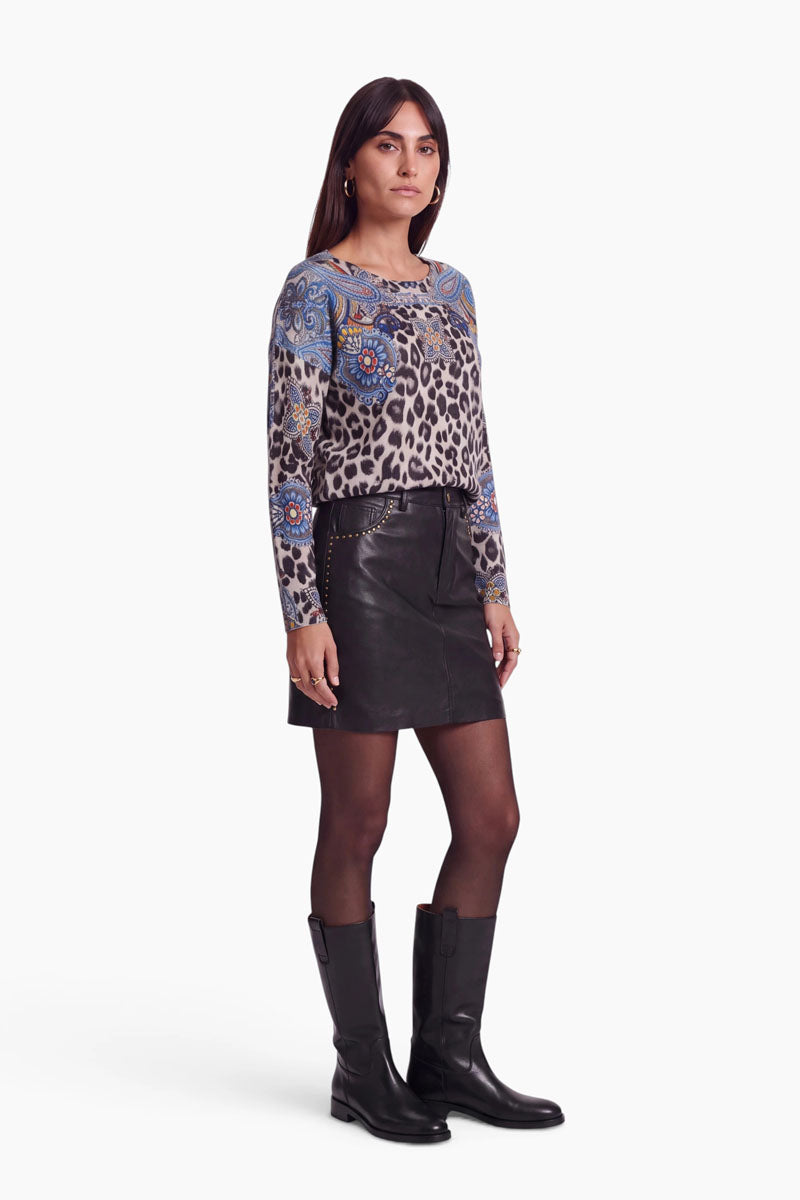 Eine Frau steht vor einem schlichten weißen Hintergrund und trägt schwarze kniehohe Stiefel, einen schwarzen Minirock, schwarze Strumpfhosen und einen langärmeligen Feinstrickpullover mit blauen Blumen- und Leo-Paisley-Mustern. Sie hat langes dunkles Haar und einen neutralen Gesichtsausdruck.