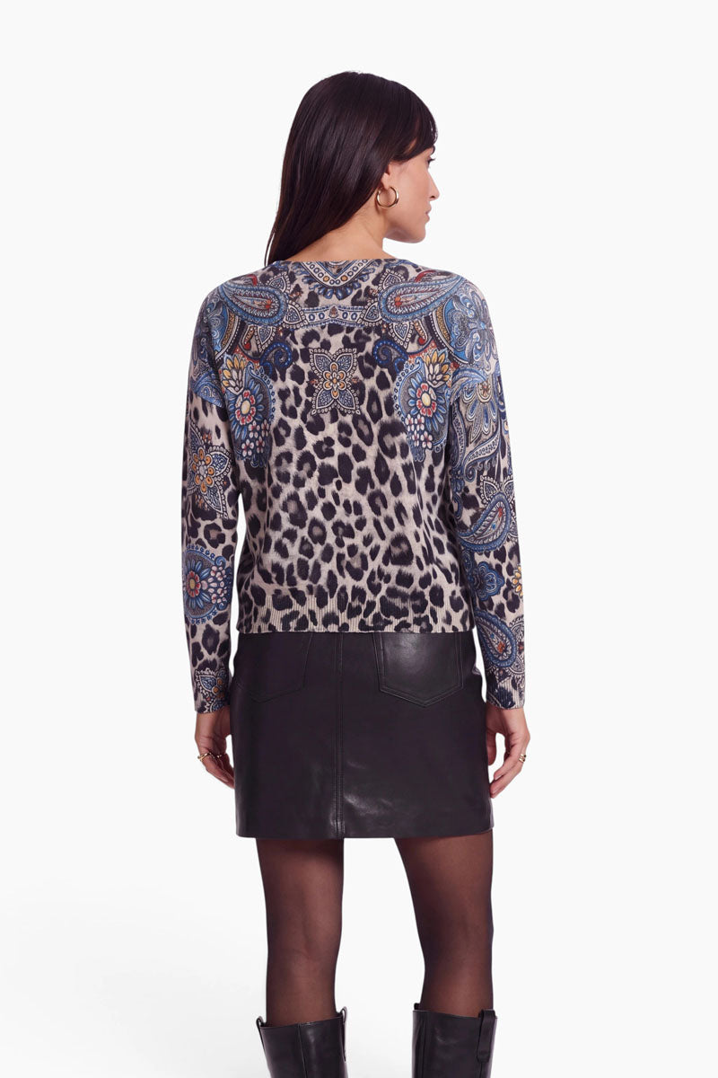 Eine Frau steht mit dem Rücken zur Kamera und trägt einen langärmeligen Feinstrickpullover mit auffälligem Leo-Paisley-Muster, dazu einen schwarzen Lederminirock, eine transparente schwarze Strumpfhose und schwarze kniehohe Stiefel. Der Pullover besteht aus einer luxuriösen Woll-Kaschmir-Mischung. Der Hintergrund ist schlicht weiß.