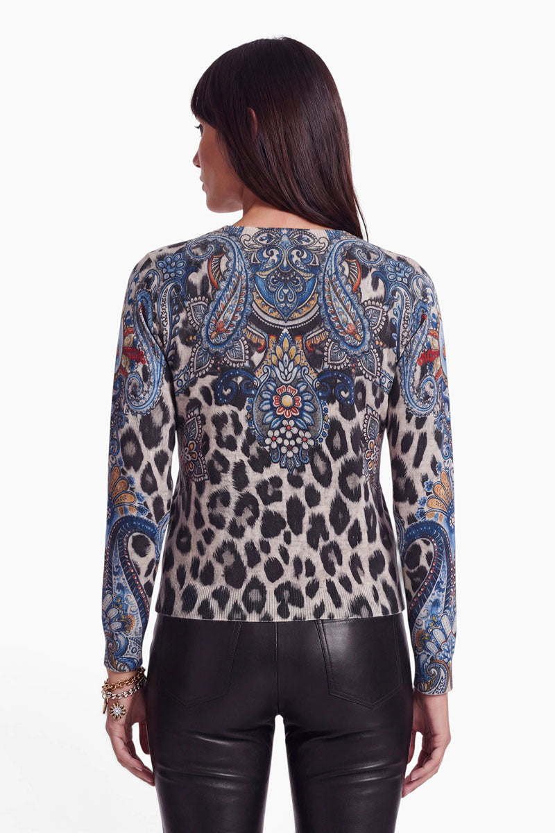 Eine Frau mit langen dunklen Haaren, von hinten gesehen, trägt eine figurbetonte Feinstrickjacke in einem auffälligen blauen Leo-Paisley-Muster, dazu eine schwarze Lederhose. Der Hintergrund ist schlicht weiß.