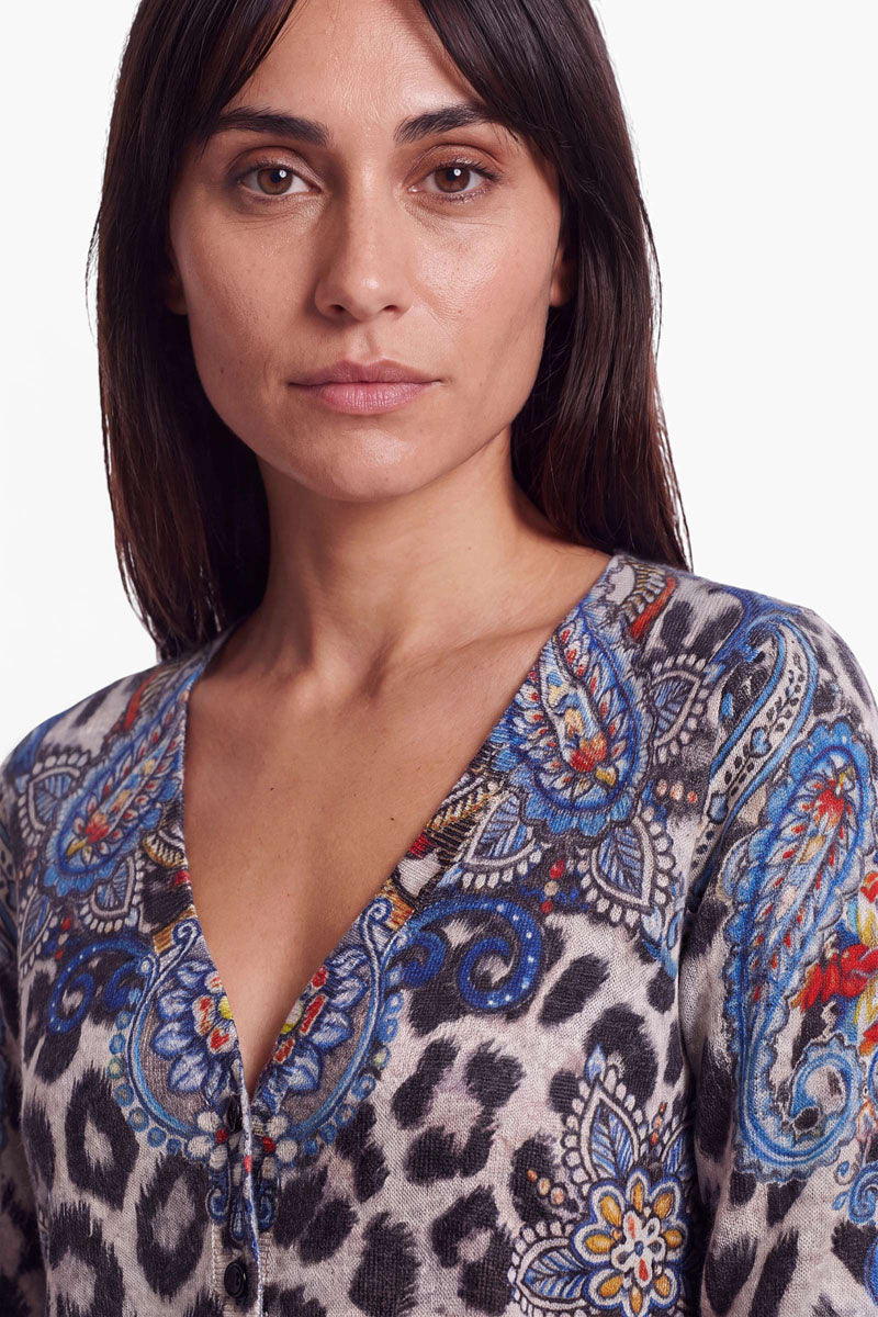Eine Frau mit glattem braunem Haar trägt eine Feinstrickjacke mit V-Ausschnitt, die durch ein auffälliges Leo-Paisley-Muster in bunten Blau-, Orange- und Gelbtönen besticht. Sie blickt mit ausdrucksloser Miene nach vorne. Der Hintergrund ist schlicht weiß.