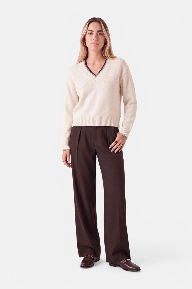 Eine Frau trägt einen cremefarbenen Pullover aus einer Woll-Kaschmir-Mischung mit V-Ausschnitt und dunklem Besatz sowie eine braune Hose mit weitem Bein. Dazu trägt sie dunkelbraune Loafer mit goldenen Beschlägen.