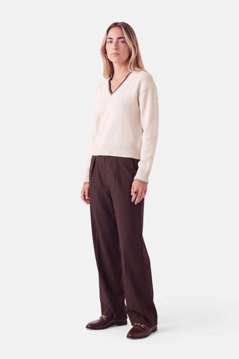 Eine Frau, die einen cremefarbenen Strickpullover mit V-Ausschnitt und braunen Bordüren sowie eine dunkelbraune Hose trägt. Sie steht und trägt braune Slipper mit goldenen Beschlägen.