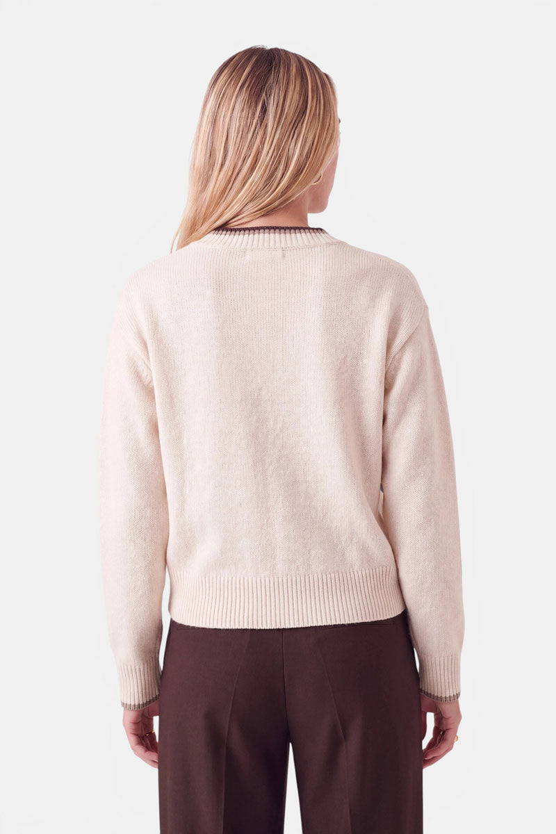 Eine Frau, von hinten gesehen, die einen cremefarbenen Pullover aus einer Woll-Kaschmir-Mischung mit V-Ausschnitt, braunen Besätzen am Ausschnitt und an den Ärmelbündchen sowie eine dunkelbraune Hose trägt.