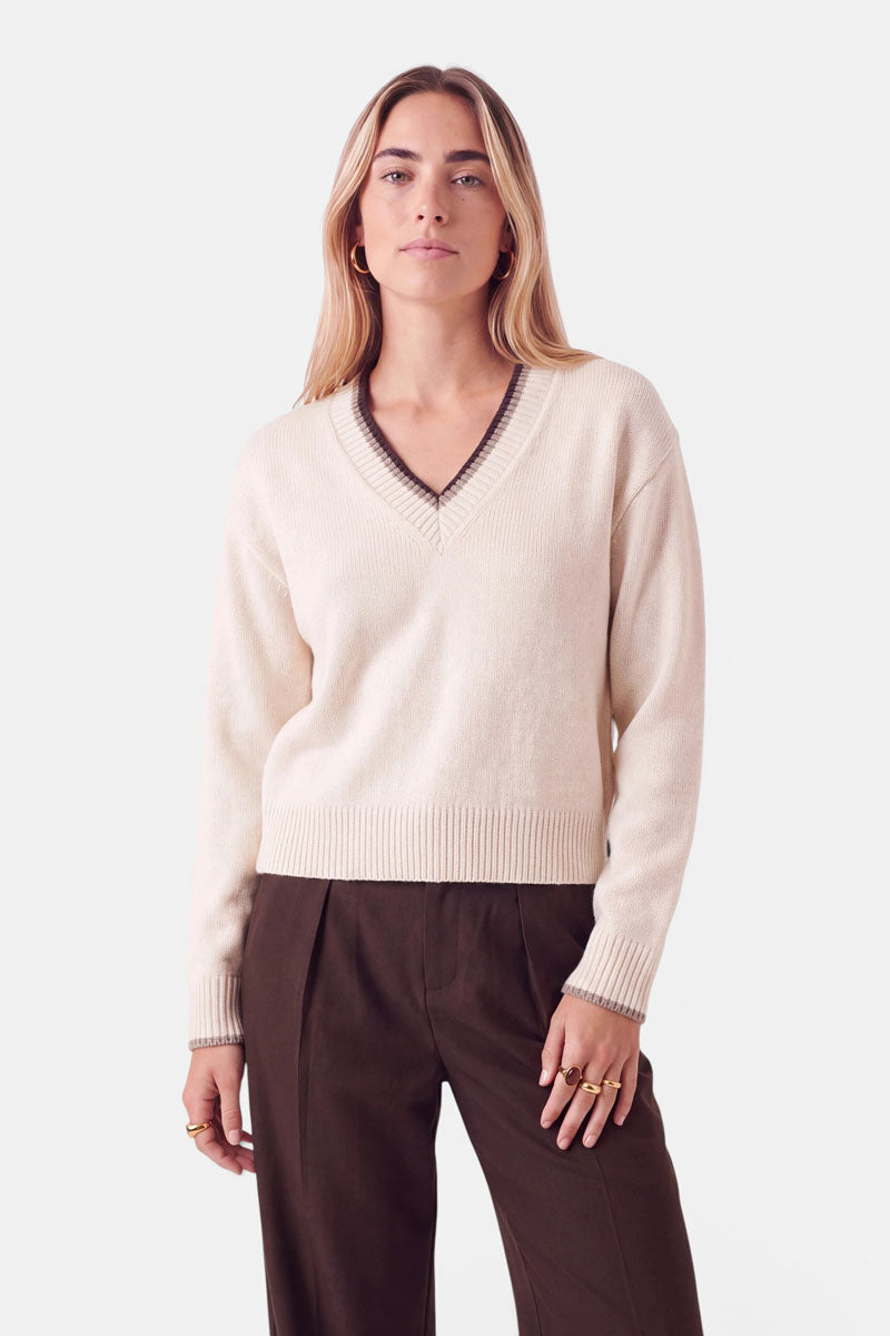Eine Frau trägt einen cremefarbenen Pullover aus einer Woll-Kaschmir-Mischung mit V-Ausschnitt, der am Ausschnitt und an den Ärmelbündchen mit dunkelbraunen Streifen verziert ist, dazu eine dunkelbraune Hose.