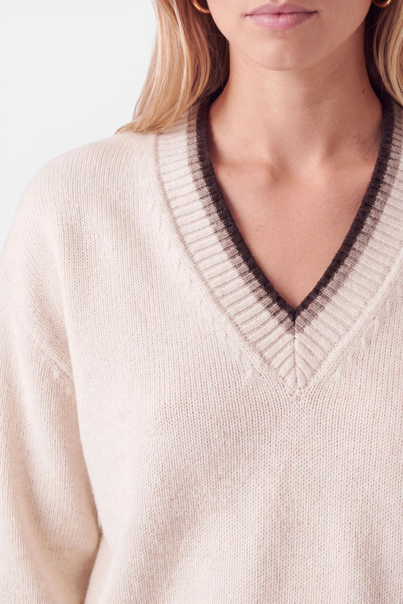 Eine Frau, die einen cremefarbenen, langärmeligen Strickpullover mit V-Ausschnitt trägt. Der Ausschnitt ist mit einem dunkelbraunen, gerippten Rand verziert.