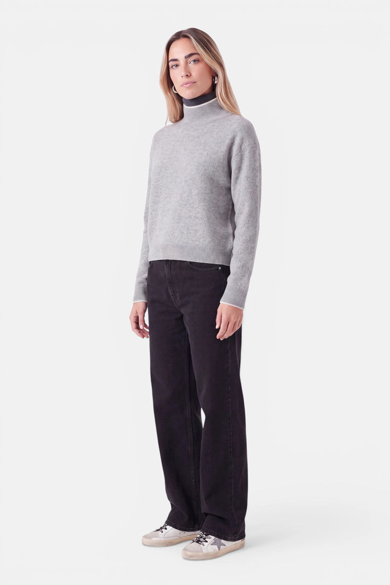 Eine Frau, die einen grauen Rollkragenpullover aus einer Woll-Kaschmir-Mischung mit weißem Rand und eine schwarze Jeans mit weitem Bein trägt. Sie steht und blickt in die Kamera; an den Füßen trägt sie Sneaker in neutralen Farben.