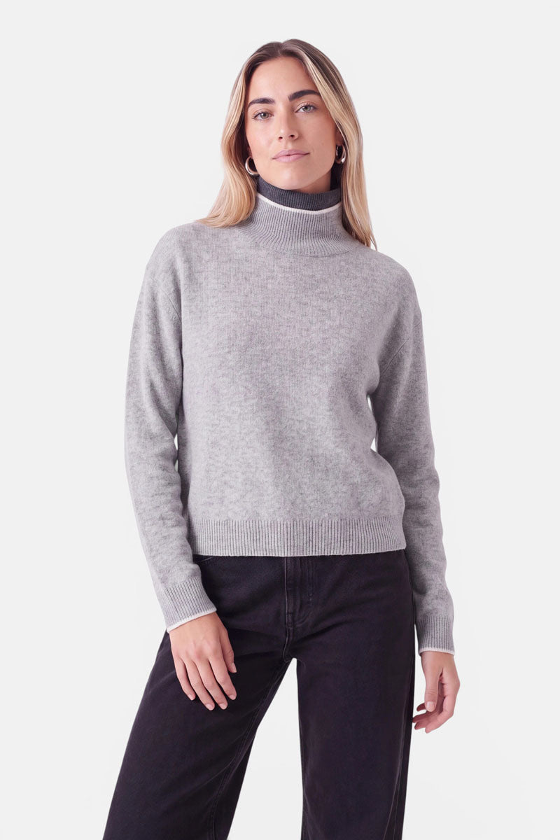 Eine Frau, die einen grauen Rollkragenpullover aus einer Woll-Kaschmir-Mischung mit dunkelgrauem Innenkragen und weißem Besatz an den Ärmelbündchen und am Saum trägt, dazu eine dunkle Jeans.