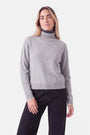 Eine Frau, die einen grauen Rollkragenpullover aus einer Woll-Kaschmir-Mischung mit dunkelgrauem Innenkragen und weißem Besatz an den Ärmelbündchen und am Saum trägt, dazu eine dunkle Jeans.