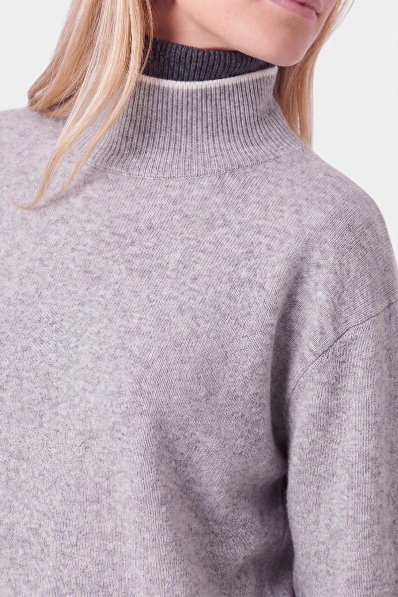 Eine Person trägt einen hellgrauen Strickpullover aus einer Woll-Kaschmir-Mischung mit geripptem Rollkragen. Der Kragen ist innen mit dunklem Anthrazit gefüttert.