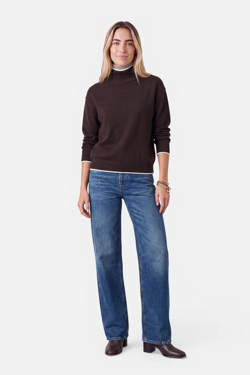 Eine Frau, die einen dunkelbraunen Rollkragenpullover aus einer Woll-Kaschmir-Mischung mit weißem Besatz am Kragen und am Saum trägt, dazu eine blaue Jeans mit geradem Bein und braune Stiefel.