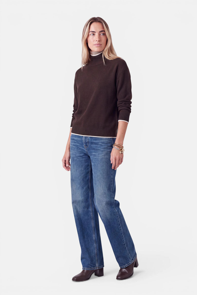 Eine Frau trägt einen braunen Rollkragenpullover aus einer Woll-Kaschmir-Mischung mit weißem Besatz am Kragen und am Saum, dazu eine locker sitzende blaue Jeans und braune Stiefel mit Absatz. Sie steht mit nach vorne gerichtetem Blick und den Armen an den Seiten.