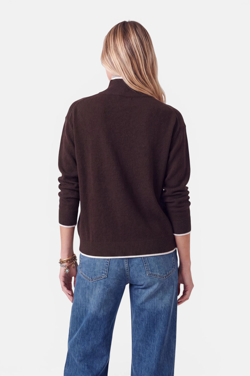Eine Person, von hinten gesehen, die einen dunkelbraunen Strickpullover mit Stehkragen und weißem Besatz am Halsausschnitt, an den Ärmelbündchen und am Saum trägt. Außerdem trägt sie eine blaue Jeans und mehrere Armbänder am linken Handgelenk.