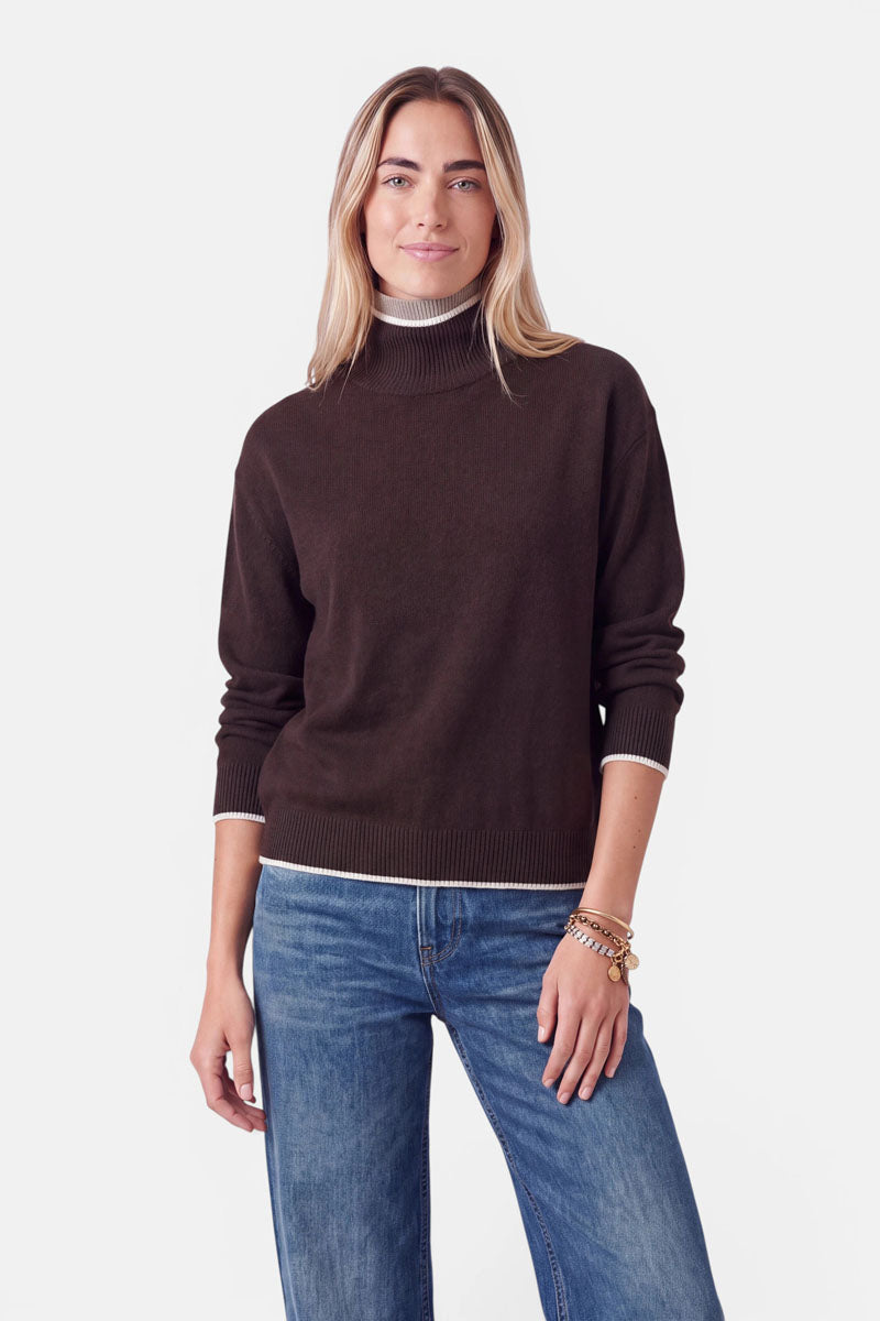 Eine Frau, die einen dunkelbraunen Rollkragenpullover aus einer Woll-Kaschmir-Mischung trägt, der am Kragen und an den Ärmelbündchen mit einem schmalen weiß-beigen Rand verziert ist. Dazu trägt sie eine blaue Jeans und mehrere Goldarmbänder übereinander.