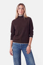 Eine Frau, die einen dunkelbraunen Rollkragenpullover aus einer Woll-Kaschmir-Mischung trägt, der am Kragen und an den Ärmelbündchen mit einem schmalen weiß-beigen Rand verziert ist. Dazu trägt sie eine blaue Jeans und mehrere Goldarmbänder übereinander.
